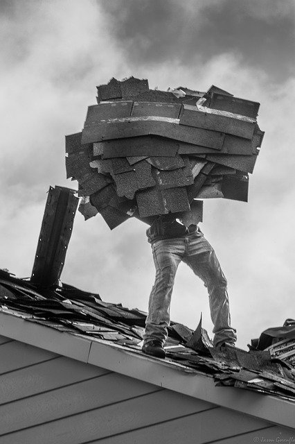 Black and white image of a man carrying pieces of roofing to be dropped off the side of the house